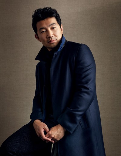 Portrait of Simu Liu wearing a dark blue coat, sitting with hands clasped in front of a neutral brown background with soft lighting.