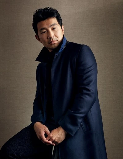 Portrait of Simu Liu wearing a dark blue coat, sitting with hands clasped in front of a neutral brown background with soft lighting.