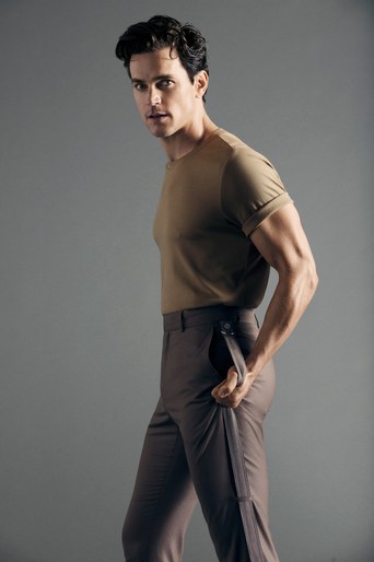 Man poses in studio, wearing khaki t-shirt and brown pants, looking over his shoulder. Neutral background, studio lighting, portrait.
