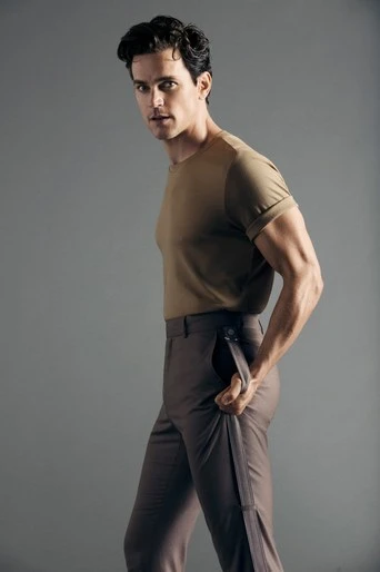 Man poses in studio, wearing khaki t-shirt and brown pants, looking over his shoulder. Neutral background, studio lighting, portrait.