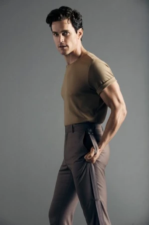 Man poses in studio, wearing khaki t-shirt and brown pants, looking over his shoulder. Neutral background, studio lighting, portrait.