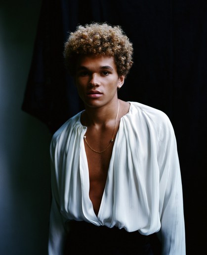 Portrait of a young adult man with curly blond hair wearing a white silk shirt in a dimly lit photo studio setting.