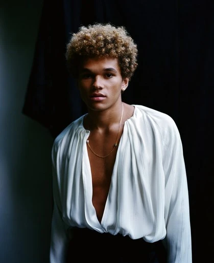 Portrait of a young adult man with curly blond hair wearing a white silk shirt in a dimly lit photo studio setting.