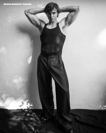 Black and white portrait of a muscular man in a tank top and pants posing with his hands behind his head against a neutral wall.