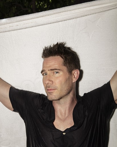 Portrait of a man in a wet black shirt, looking at the camera. He is holding a white textured fabric above his head.
