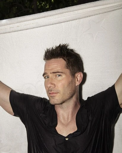 Portrait of a man in a wet black shirt, looking at the camera. He is holding a white textured fabric above his head.