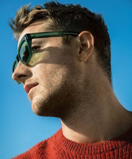 Close-up portrait of a blonde man with green sunglasses looking to the left against a bright blue sky, wearing a red sweater.