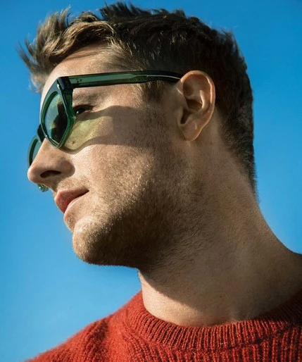 Close-up portrait of a blonde man with green sunglasses looking to the left against a bright blue sky, wearing a red sweater.