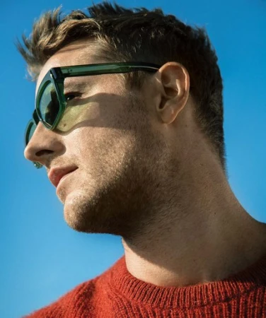 Close-up portrait of a blonde man with green sunglasses looking to the left against a bright blue sky, wearing a red sweater.