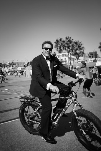 Monochrome shot of a man in a suit riding an electric bicycle on a paved area with palm trees and other people in the background.