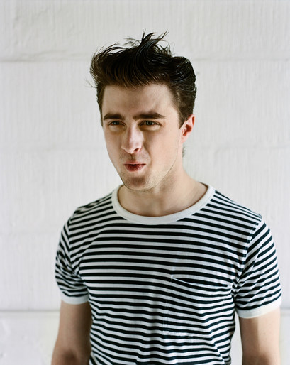 Portrait of a young adult male with dark hair wearing a striped shirt against a white brick wall backdrop indoors.