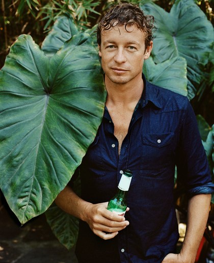 Adult male poses with a green beer bottle near a large leaf. He wears a dark-blue denim shirt in an outdoor setting.