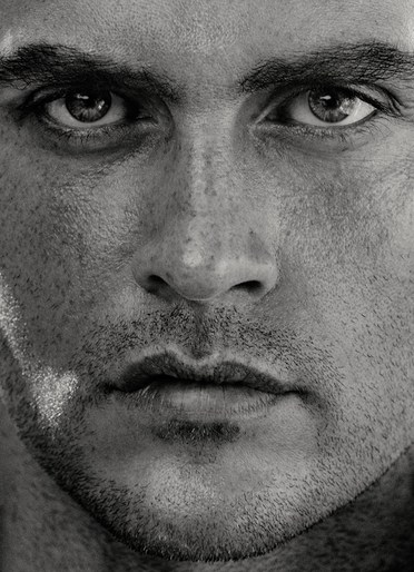Close-up portrait of a man with intense gaze, highlighting facial features, skin texture, and detail in a high-contrast, black and white image.