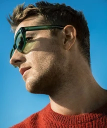 Profile portrait of a man wearing green sunglasses and an orange knit sweater against a bright blue sky.