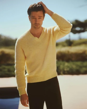 Portrait of an adult male in a yellow sweater with his hand in his hair, set against a blurred outdoor background.