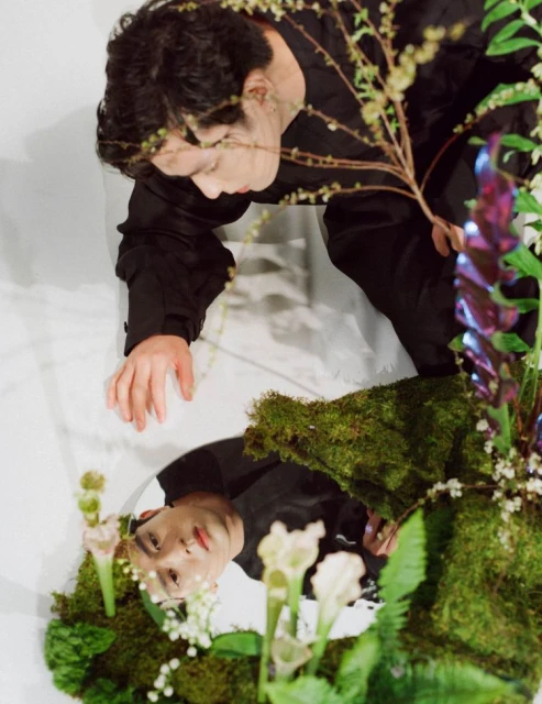 Person in black jacket crouching in front of mirror surrounded by flowers and moss, reflecting their face and greenery.