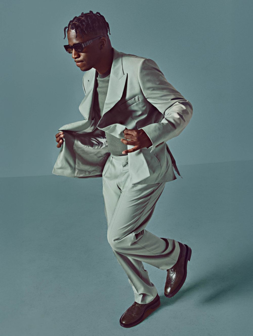 A black man in a light grey suit strikes a pose with sunglasses and brown shoes, set against a matching grey backdrop.