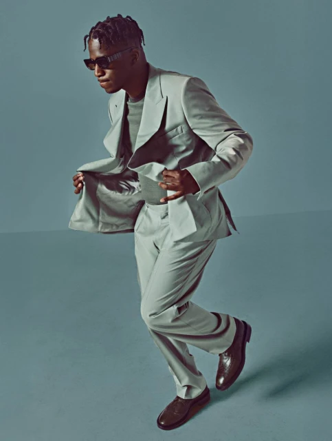 A black man in a light grey suit strikes a pose with sunglasses and brown shoes, set against a matching grey backdrop.