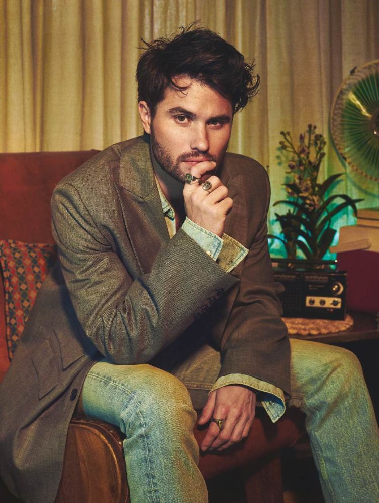 Portrait of a young adult male with dark hair wearing a khaki blazer, jeans, and rings sitting indoors in front of a fan.