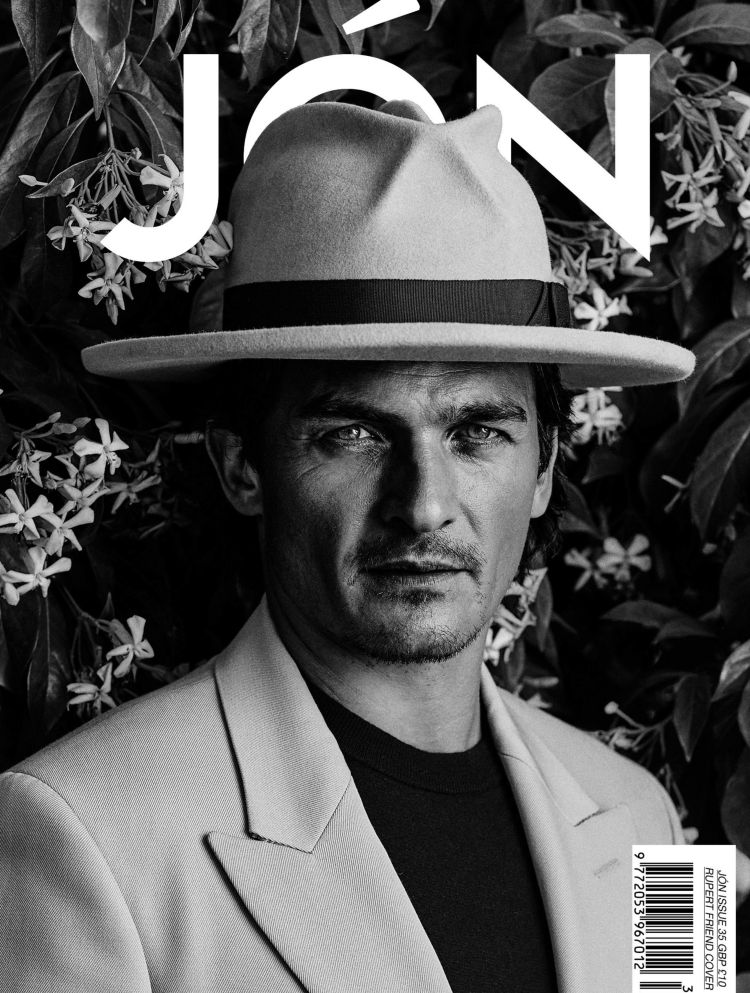 Black and white magazine cover portrait of an adult male wearing a hat and suit jacket, with a floral background.