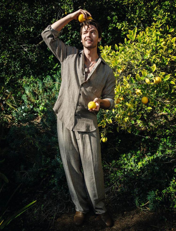 Full-body portrait of a man posing in a garden with lemons, wearing a linen suit and holding a lemon above his head.