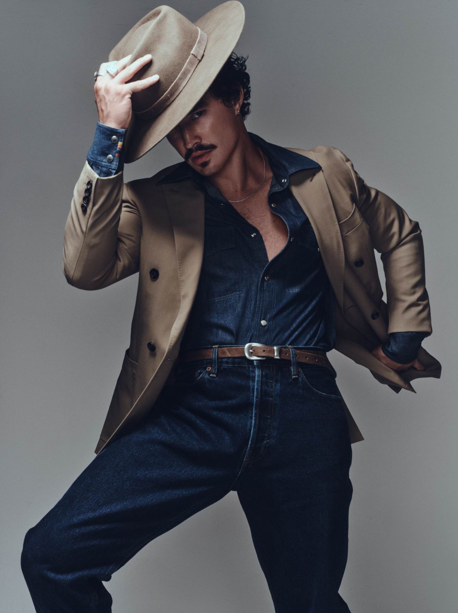 Man in a tan hat and double-breasted blazer poses in front of a grey backdrop, looking down with a slight smile in a fashion shot.