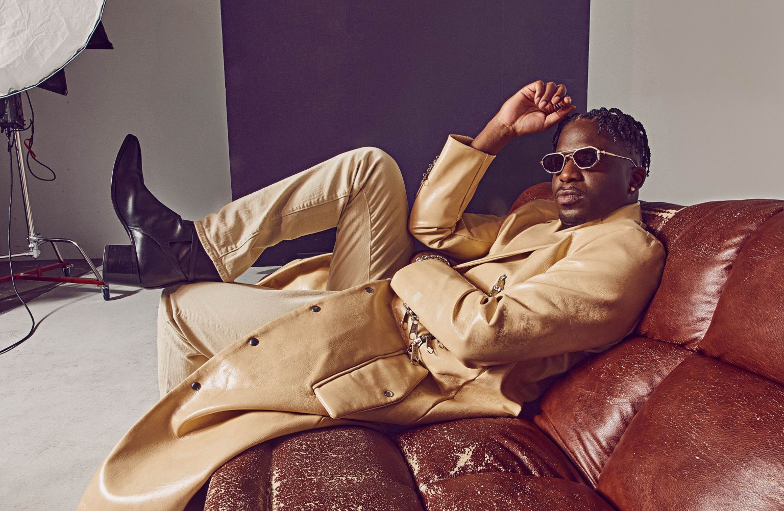 Stylish adult male in sunglasses lounges on a brown couch in a photo studio wearing a beige overcoat, pants, and black boots.