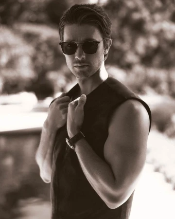 Man wearing sunglasses and a sleeveless vest, holding his collar outdoors in a portrait styled by Avo Yermagyan.
