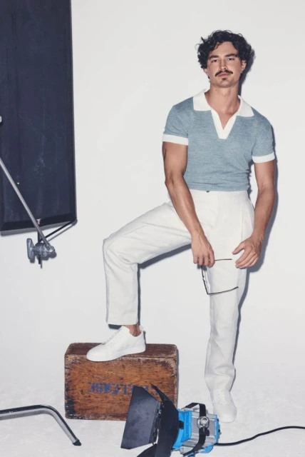 Stylish man in a studio wearing a blue polo shirt and white pants, posing next to lighting equipment and a vintage wooden crate.