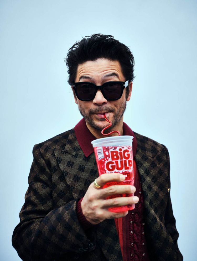 Adult male wearing sunglasses and a patterned jacket drinking a red soda from a Big Gulp cup with a red straw against a light blue backdrop.