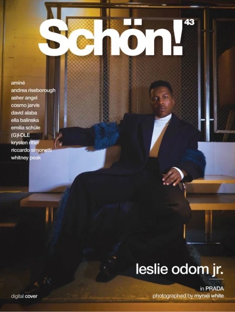 Leslie Odom Jr. is sitting, posing on a bench and wearing a black suit with fur sleeves for Schön! magazine digital cover.