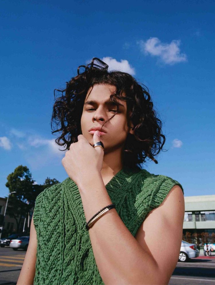 Young man poses in a green knitted vest with blue sky and urban street background. Bracelet and ring visible. Shoulder-length curly hair.