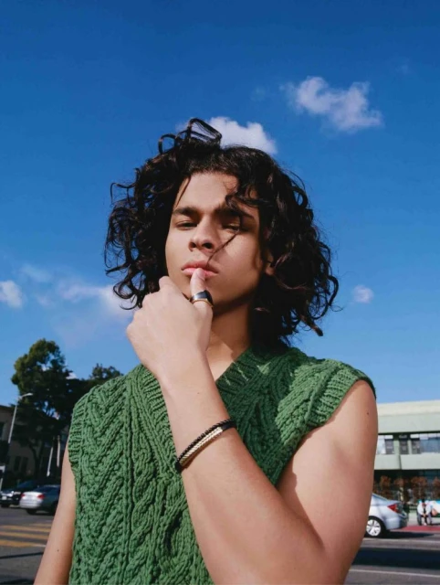 Young man poses in a green knitted vest with blue sky and urban street background. Bracelet and ring visible. Shoulder-length curly hair.