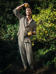 Man wearing a grey linen shirt and trousers holding fresh lemons in a sunlit orchard, styled by Avo Yermagyan.