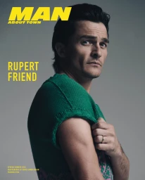 Portrait of Rupert Friend wearing a green knit sweater against a grey studio backdrop, styled by Avo Yermagyan.