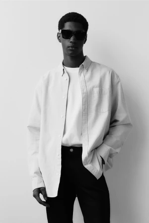 Black and white studio portrait of a young Black man wearing sunglasses, an unbuttoned shirt over a t-shirt, and dark trousers.
