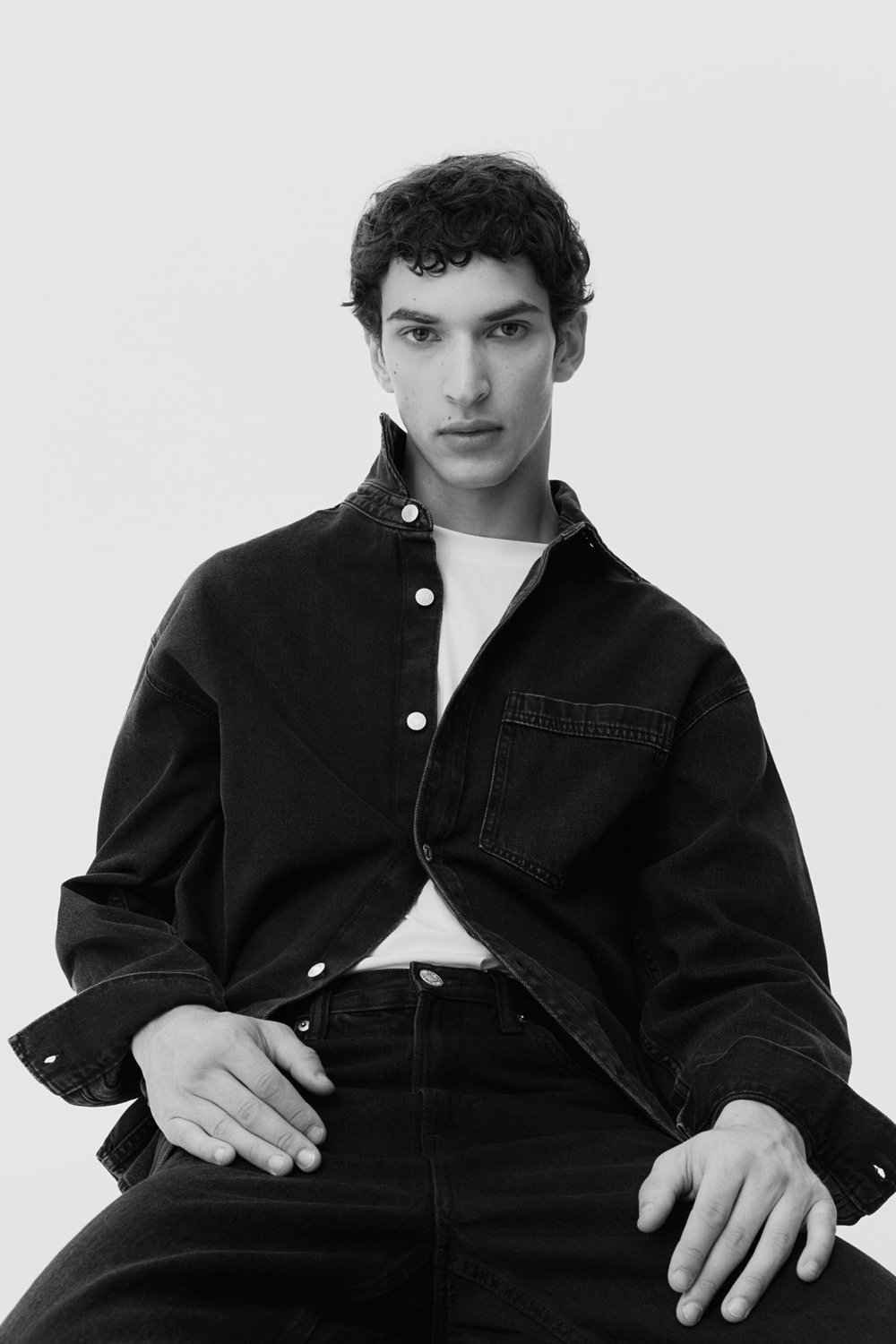Black-and-white portrait of a dark-haired male model sitting, wearing a denim jacket and jeans against a light background in a studio setting.