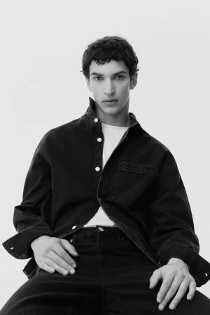 Young man in a dark denim shirt and white t-shirt posing against a plain light background styled by Alberto Vegue.