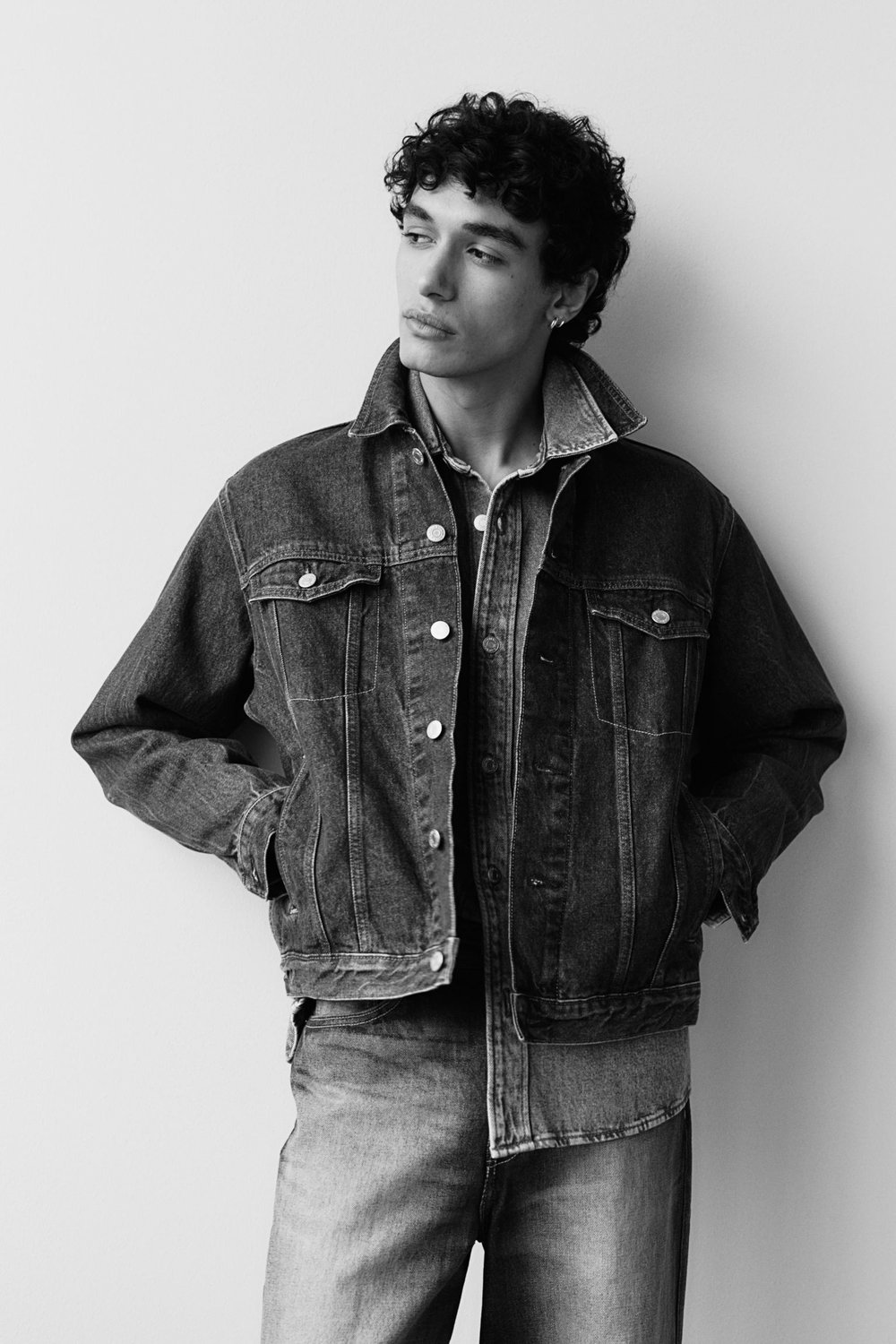 Black and white portrait of a male model in denim jacket and jeans against a neutral backdrop in a fashion studio.