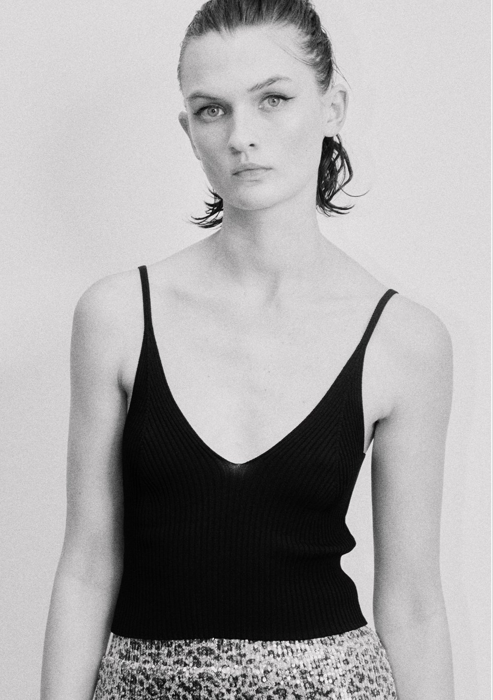 Black and white portrait of a woman with fair skin wearing a black V-neck top and patterned skirt, posing against a light grey background.