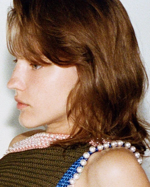 Close-up studio shot of a woman with wavy brown hair and a light complexion, wearing a dark green top and beaded jewelry.