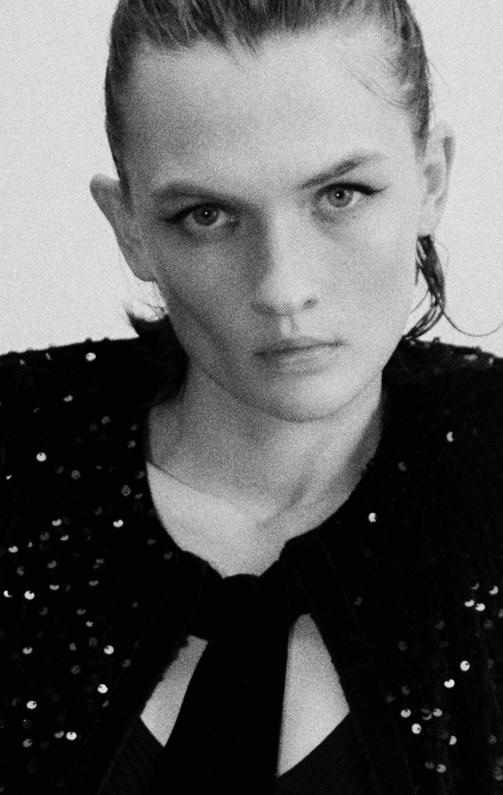 Monochrome portrait of a young woman with dark hair, looking directly at the camera, wearing a sequined top against a plain backdrop.