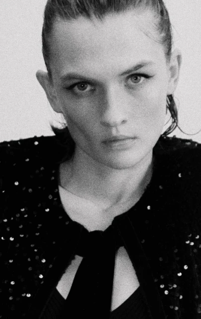Monochrome portrait of a young woman with dark hair, looking directly at the camera, wearing a sequined top against a plain backdrop.
