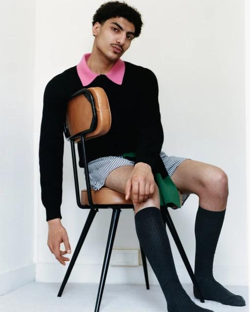 Young man with dark hair and a mustache sits on a chair in a studio, wearing a sweater, shorts, and knee-high socks in a simple indoor setting.