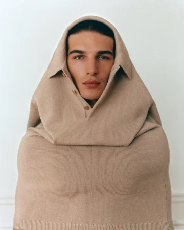 Close-up portrait of a man wearing a beige knit polo shirt pulled over his head like a hood, styled by Alberto Vegue