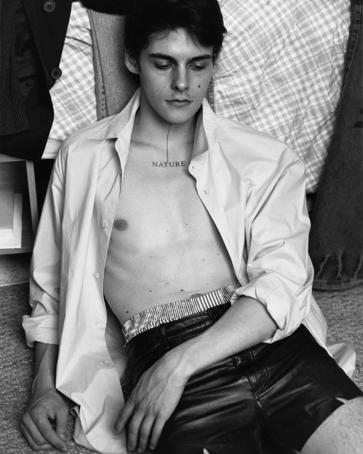 Black-and-white portrait of a young adult male lying down, wearing a partly unbuttoned white shirt and black leather shorts indoors.