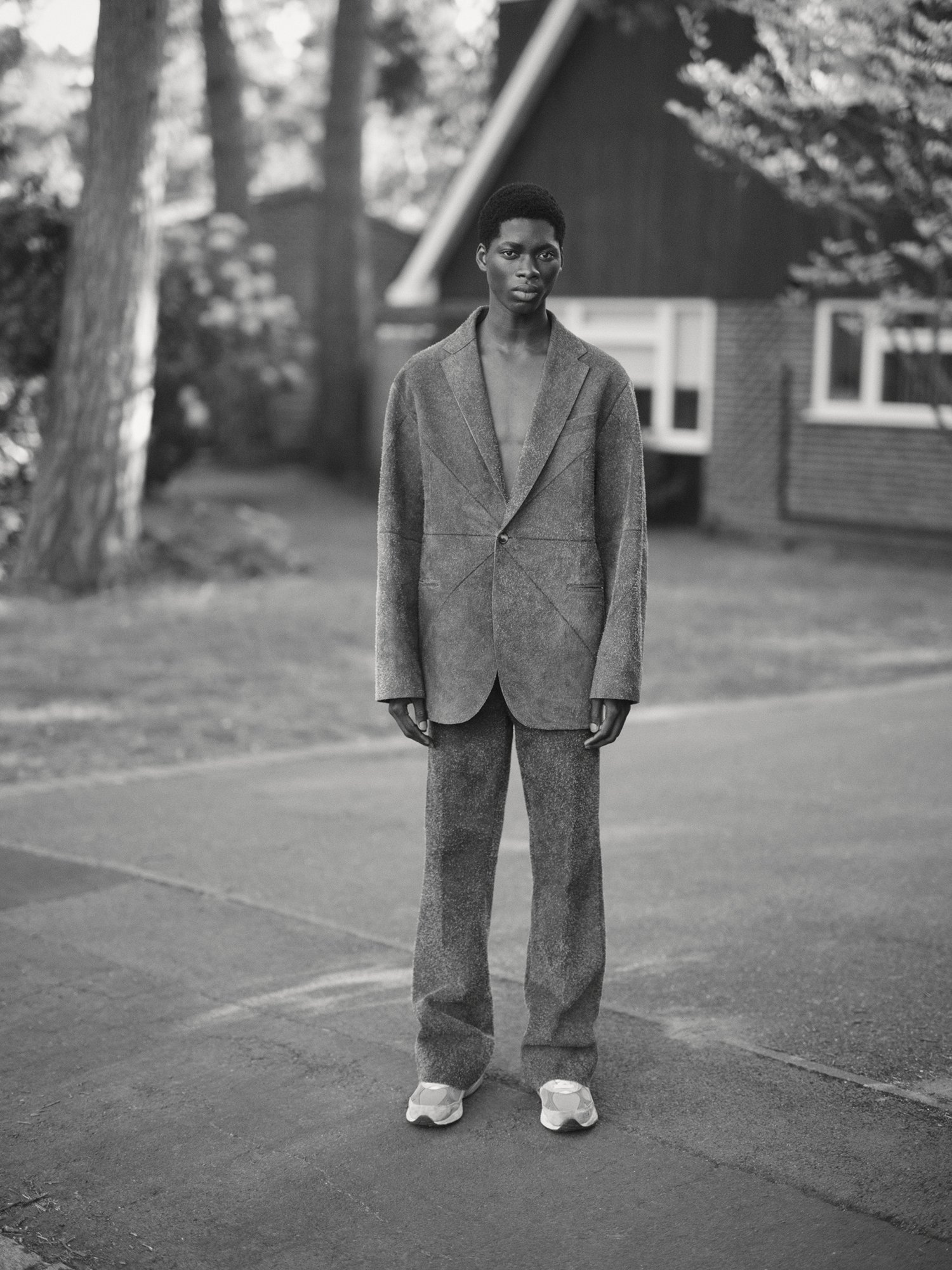 Monochrome portrait of a Black man in a suit standing outside a house, wearing sneakers, with trees and greenery in the background.