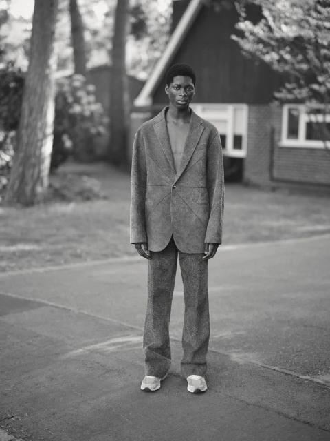 Monochrome portrait of a Black man in a suit standing outside a house, wearing sneakers, with trees and greenery in the background.