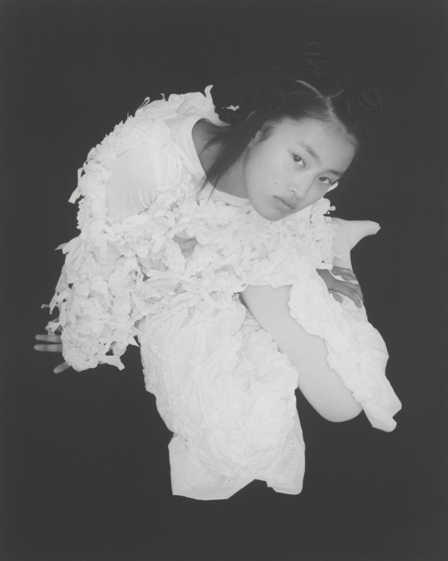 Monochrome portrait of an Asian female model in an abstract white dress against a black background, shot from a high angle.