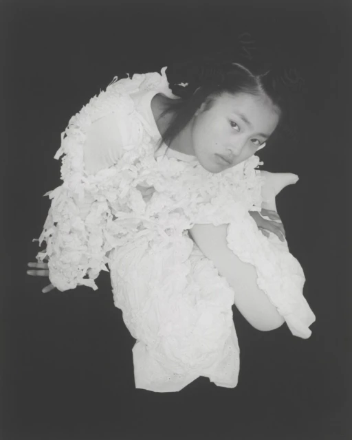 Monochrome portrait of an Asian female model in an abstract white dress against a black background, shot from a high angle.